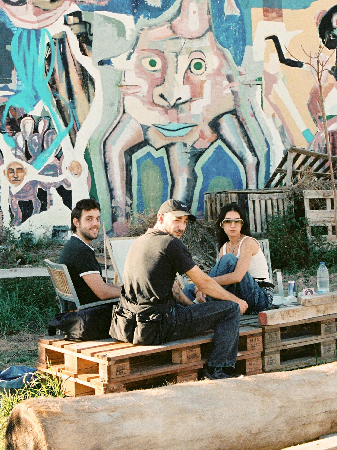 Tres personas sentadas sobre palés frente al gran mural pintado en la fachada de las naves de Pluto